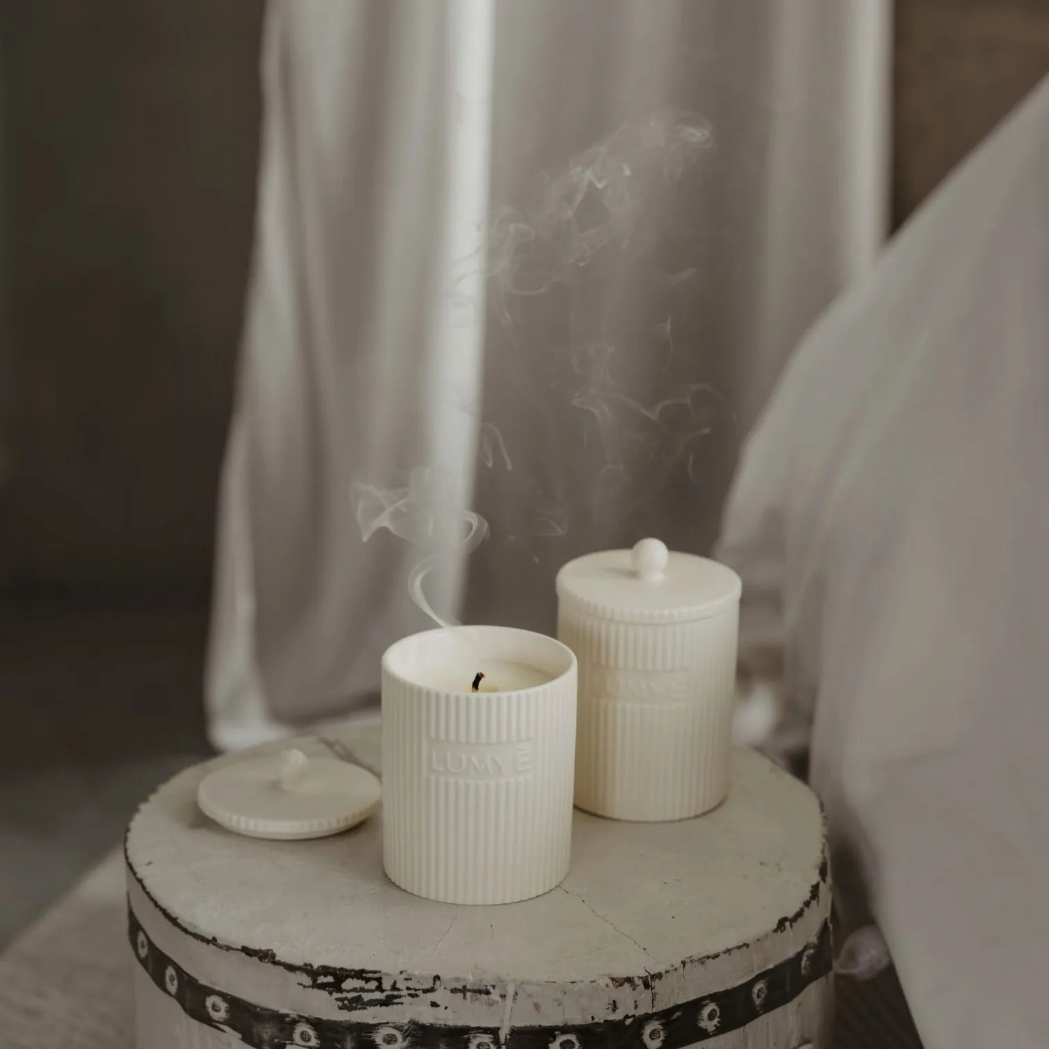 Dwie świece LUMYÈ SAONA w kremowej ceramice z unoszącym się dymem obok łóżka